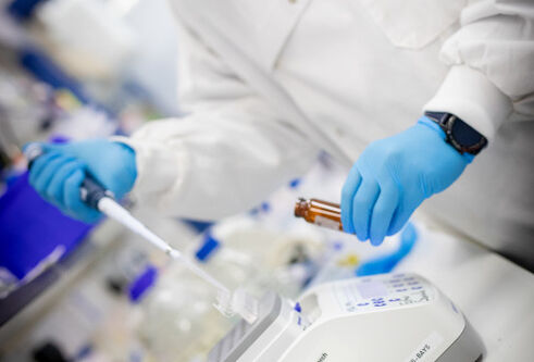 A person pipetting into a brown vial wearing a lab coat and blue gloves
