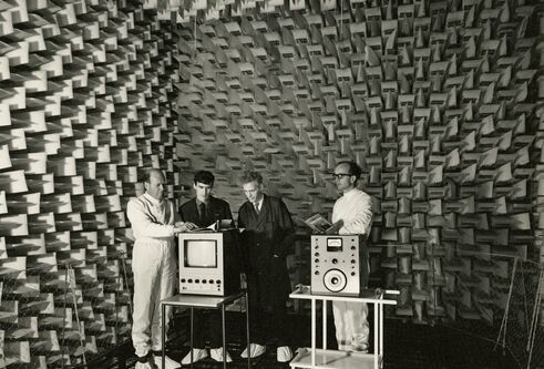 Four men stand inside an Anechoic Chamber. They are looking at two pieces of equipment, one a monitor, the other some sort of measurement device