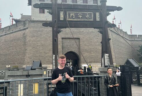 A man in a black tshirt and blue jeans stands in front of a Chinese building with two thumbs up