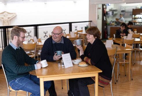 Three people sit around a wooden table smiling and drinking from mugs.