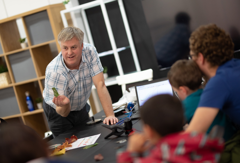 Man crouched over a table and laptop holding up a leaf and looking towards a man and two small children, who have their backs to us