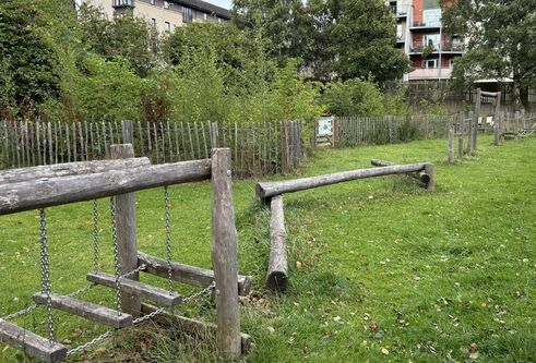 Kids wooden playframe and climbing equipment in grassy are with trees surrounding and tall buildings behind 