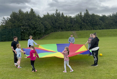 A group of adults and children stand in a circle holding the edges of a rainbow coloured parachute, in a field with trees in the background.