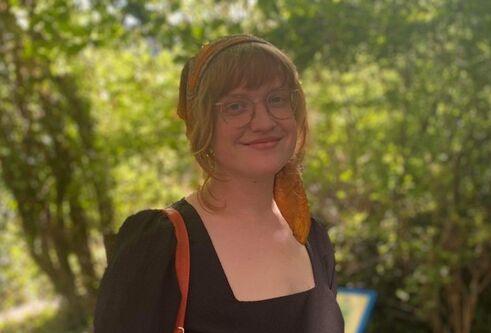 woman standing in forest looks towards camera and smiles. She is wearing a black top with a read bag strap over her shoulder, glasses and has fair hair tied back and a fringe 