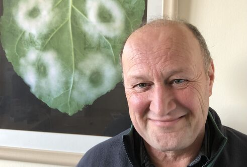 Head and shoulders shot of a man smiling. He is in front of a framed image of underside of green heart shaped leaf which has 4 fluffy circles, two on each side of the leaf.