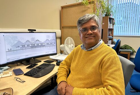 man wearing yellow woollen jumper and glasses, looking and smiling at camera. He is sat next to desk with a computer at it and a sketch of a building on the screen