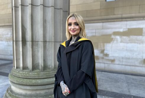 Young woman with long, straight blond hair and tattoos wearing a graduation gown, looking and smiling at camera