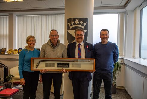 Four people stand smiling, holding a trophy case containing a putter and 3 plaques with names engraved.