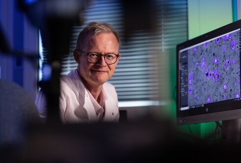 Man wearing glasses and whit lab coat looking towards the camera. Next to him is a screen with science cells on it. the image features blue and green lighting