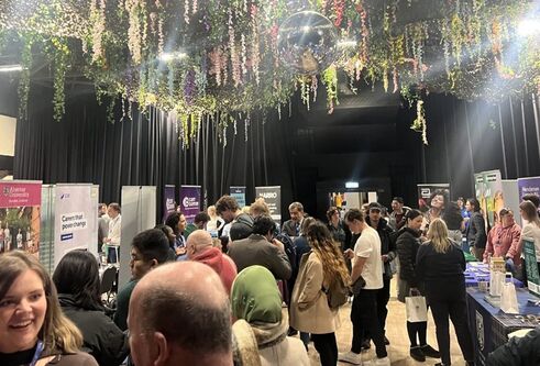 A crowd of people are in a room with flowers hanging from the ceiling, there are tables with banners promoting employers.