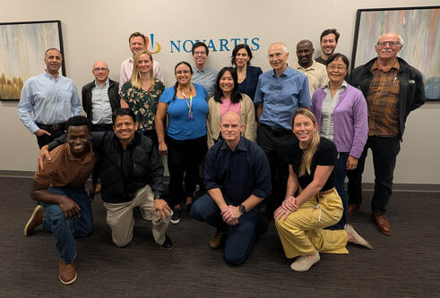 A group of people standing and kneeling together smiling in front of a sign saying Novartis