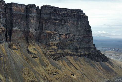 Aerial view of a steep cliff with layered rock formations and a flat landscape stretching into the distance under a cloudy sky.