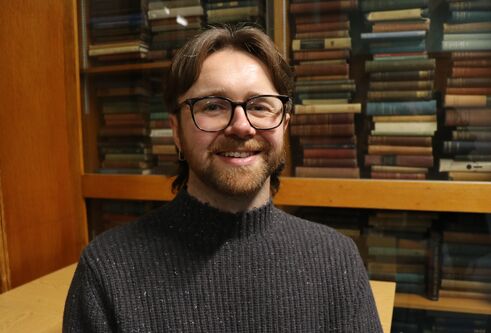 man with mid length dark hair sat in front of bookcase filled with books, looking and smiling at camera