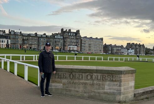A man stands in front of a wall at the Old Course golf course, in St Andrews, there is a big stone building in the background.