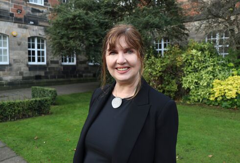 Woman with tied back dark hair, waring black smart outfit, looking and smiling at camera. She is outside in a garden area, with green bushes and grass behind her and old buildings