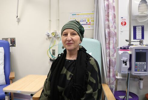 woman sat upright in chair, with green top and hat on, looking and smiling at camera. She is in a hospital room with some medical equipment around her