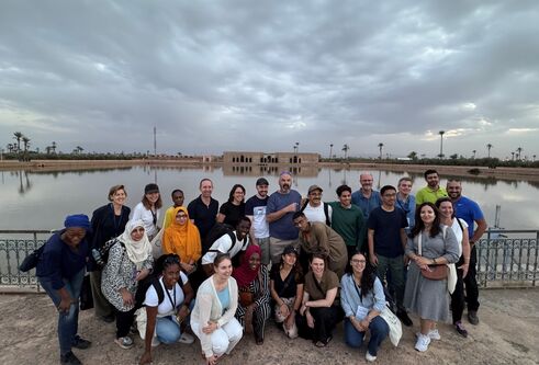 A group of people pose in front of a body of water with a building on the opposite shore.