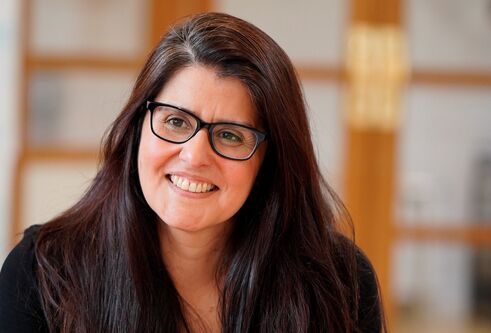 Close up head and shoulders shot of woman with long dark hair and glasses, looking slightly off camera and smiling