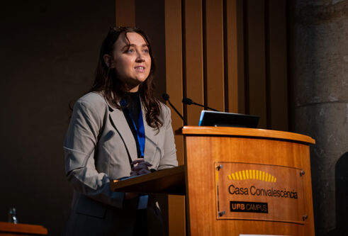 A girl with brown hair, wearing a grey blazer stands at a podium looking out to the top right.