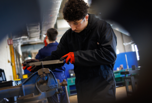 A student in a boiler suit and gloves works a piece of metal being held in a vice