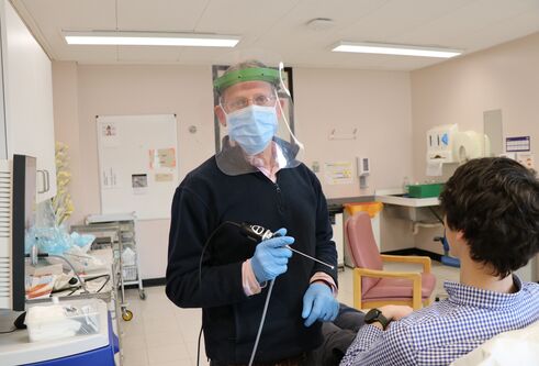 man wearing a face mask and protective clear facial visor, holding a long thin tool with a light on the end of it. He is looking towards the camera. A patient is seen from behind lying on a hospital bed.