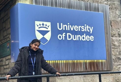 A woman in black clothing leans against metal railing outside of a building, there is a large blue 'University of Dundee' sign behind her.