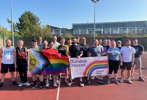 A crowd of people stand on an outdoor tennis court, holding LGBTQ+ and the Dundee Deuces flag.