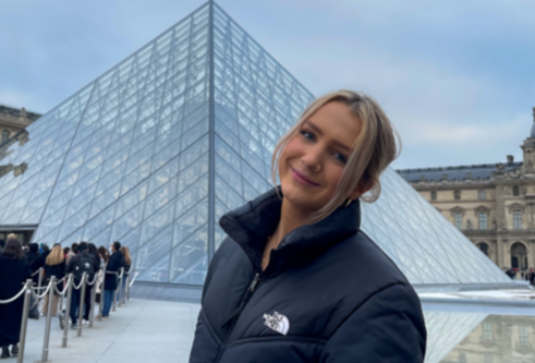 A blonde woman in a black coat is in front of the louvre.