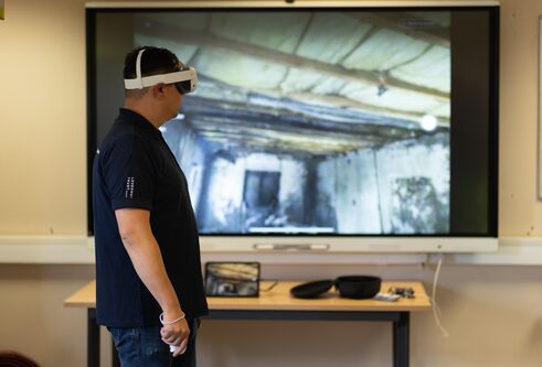 Man wearing a VR headset with a large screen behind him with showing a burnt out room