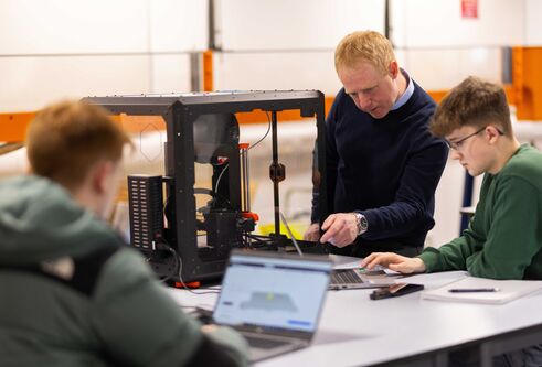 Two students work on laptops next to a 3d printing machine, a tutor stands next to one of the students pointing at a laptop screen.