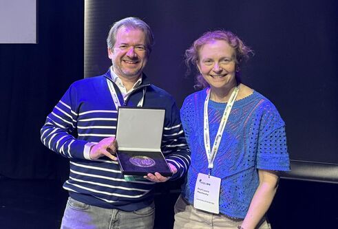Man and woman standing next to each other both smiling and looking at the camera. Man is holding a large medal in an open box.