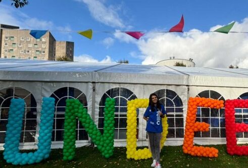 A person stands in front of a white marquee with colourful bunting flying overhead. There is large writing spelling out "DUNDEE" with balloons.