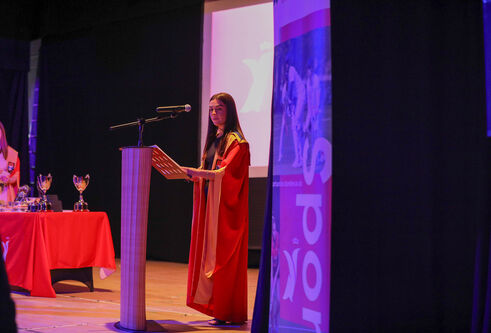 A person in a robe stands in front of a pedestal speaking in a microphone.