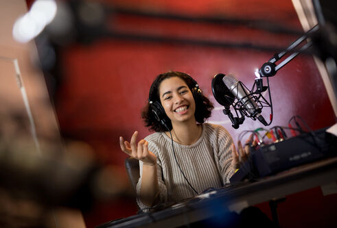 A person sits at a microphone wearing a headset and smiling with their hands raised in front of them