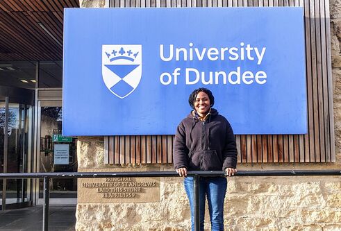 A woman stands with her hands on black metal railing, in front of a blue University of Dundee sign.