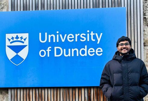 A person stands in front of a large blue University of Dundee sign, with his hands in his jacket pockets, smiling.