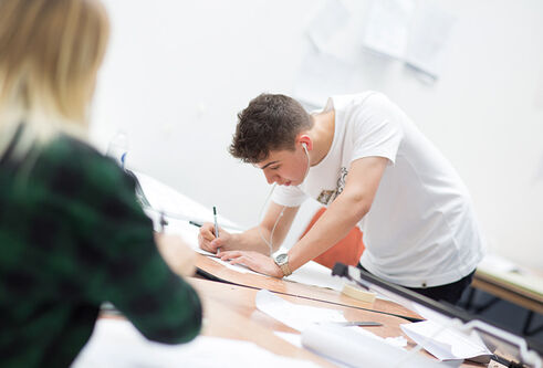 A male in a white t-shirt leans over a desk with a pencil in his hand, drawing.