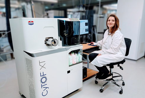 A person in lab coat sat on a chair in front of a desktop computer. Attached to the computer is a large box approx. 1.5m high which contains the scientific instrument.