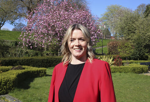 Kirsty Thomson wearing a bright red blazer, standing in a well tended garden with a pink blossoming tree behind her.
