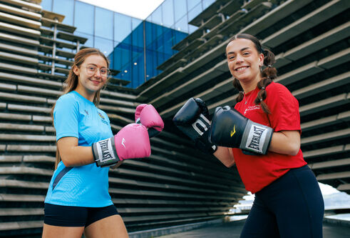 Two people stand in front of the V&A building holding their hands up to each other, wearing boxing gloves.
