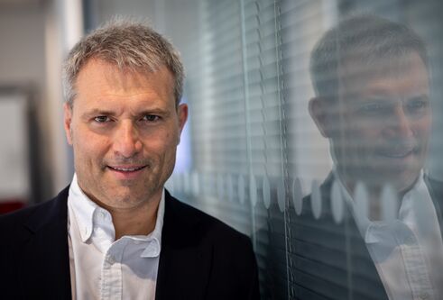 Man in white shirt and dark jacket looking at camera with his reflection seen in the glass wall on his left.