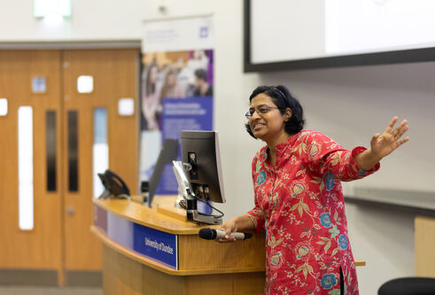 Dr Edzia Carvalho addresses visitors, standing infront of a podium in a lecture theatre.