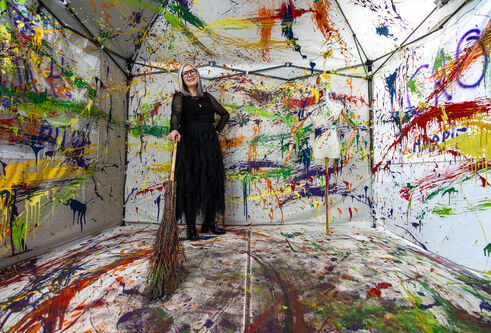 Nicky standing in her installation, holding a witches' broom. The walls are cover in different colours of paint and a mannequin is in the foreground, also covered in differnet colours.