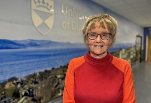 Professor Jill Belch, in a red jumper, standing in front of a blue background with the University of Dundee branding