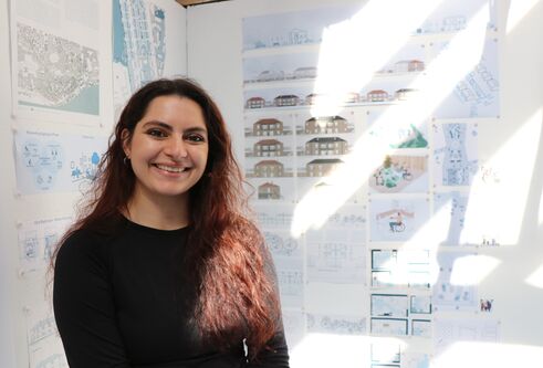 Young woman with long dark hair looking at camera and smiling, with architecture drawings on a wall behind her