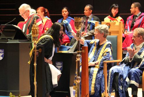 Anju Matthew on stage at a graduation ceremony, getting bopped on the head with a scroll. People in ceremonial robes are sitting.