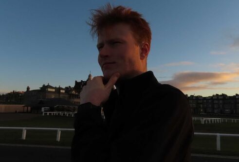 Jarret Fiske with his hand on his chin, looking thoughtful, there is grass behind with white fencing and buildings.