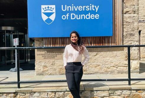 Thusharika standing in front of a large blue University of Dundee sign