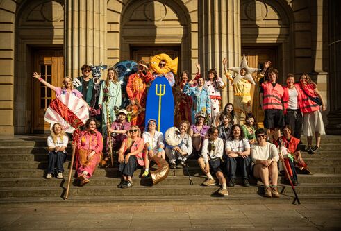 A large, costumed group poses on the steps of Caird Hall in the sunshine, many dressed in colourful handmade outfits.