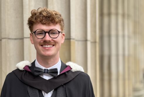 Graduate Kieran, wearing a graduation robe and standing on Caird Hall steps